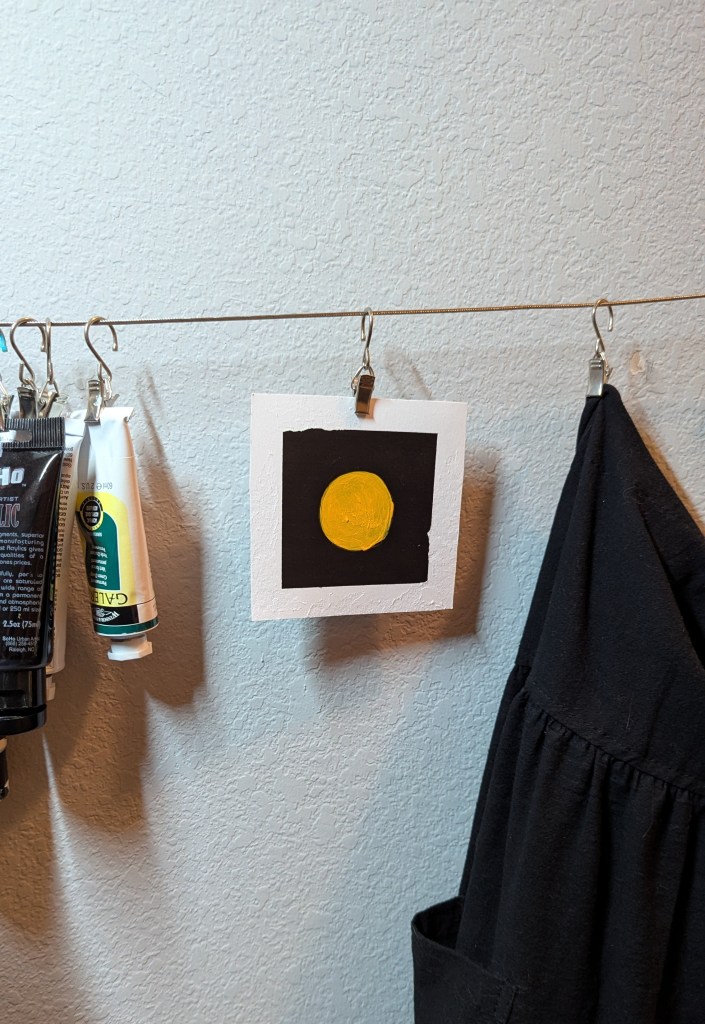 A paper square with a black background and a textured, handpainted orange circle on it. It hangs on a wire, with an apron and paint tubes hanging on either side of it.